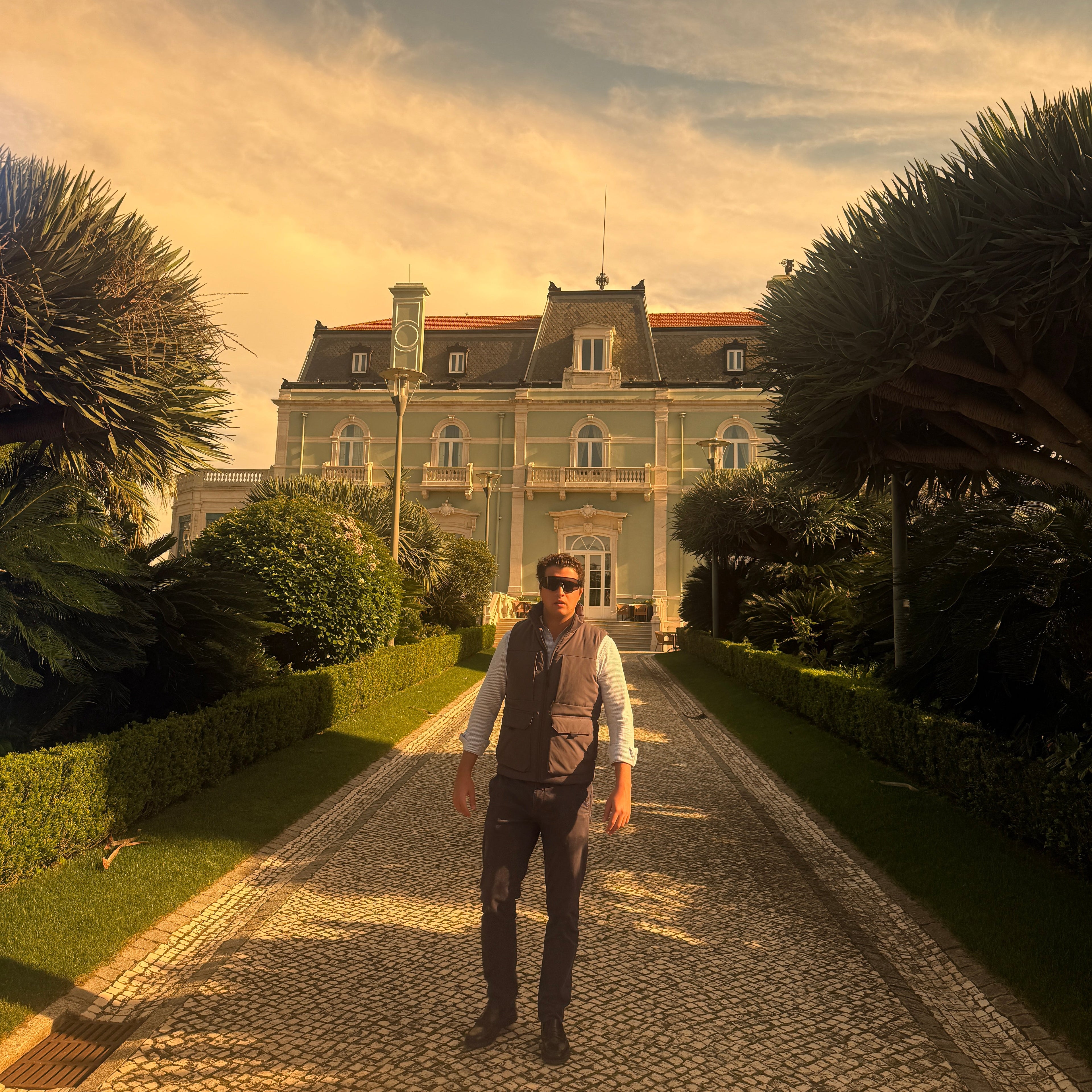Person standing on a cobblestone path in front of a large building with palm trees.