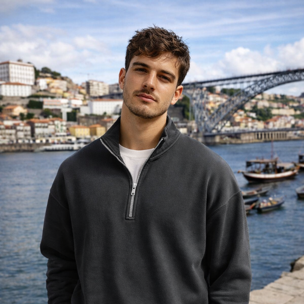 Man wearing a dark gray pullover with a zipper, standing by a waterfront with a bridge and buildings in the background.