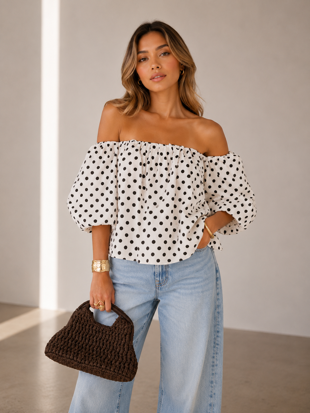 Woman wearing a polka dot top and jeans, holding a brown handbag indoors.