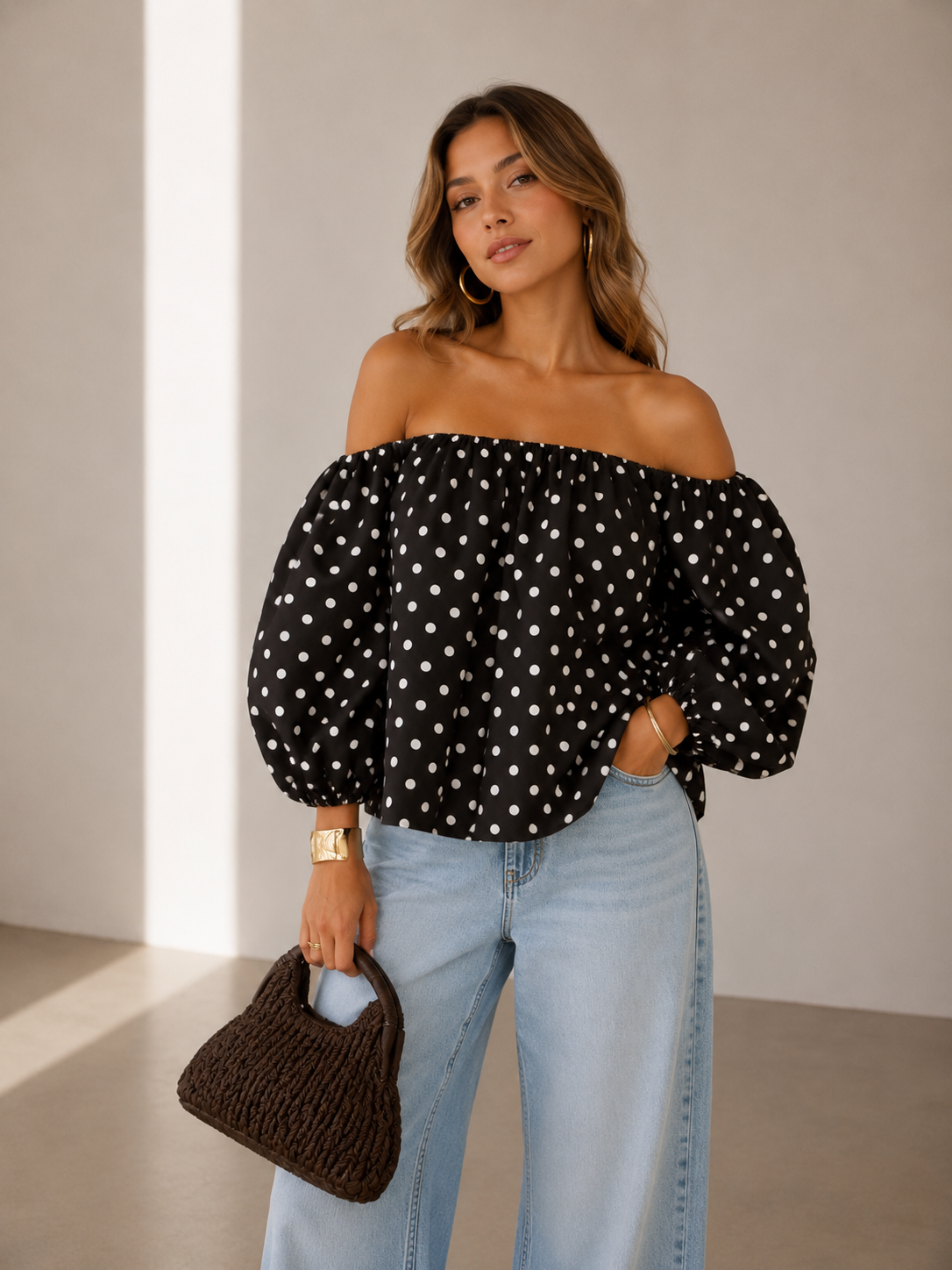 Woman wearing a black off-shoulder top with white polka dots and light blue jeans, holding a brown handbag.