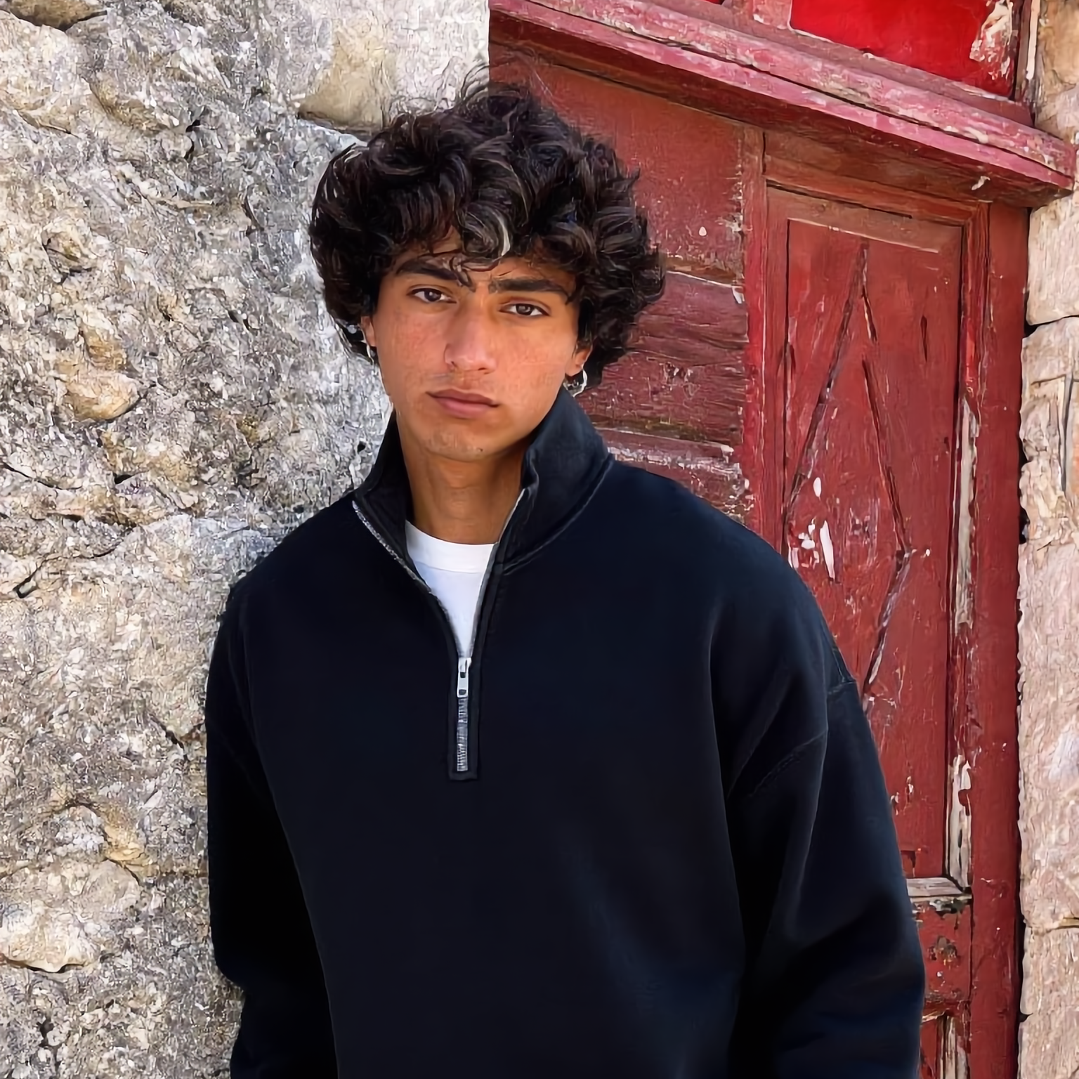 Person wearing a black sweater and light pants standing against a stone wall with a red door.