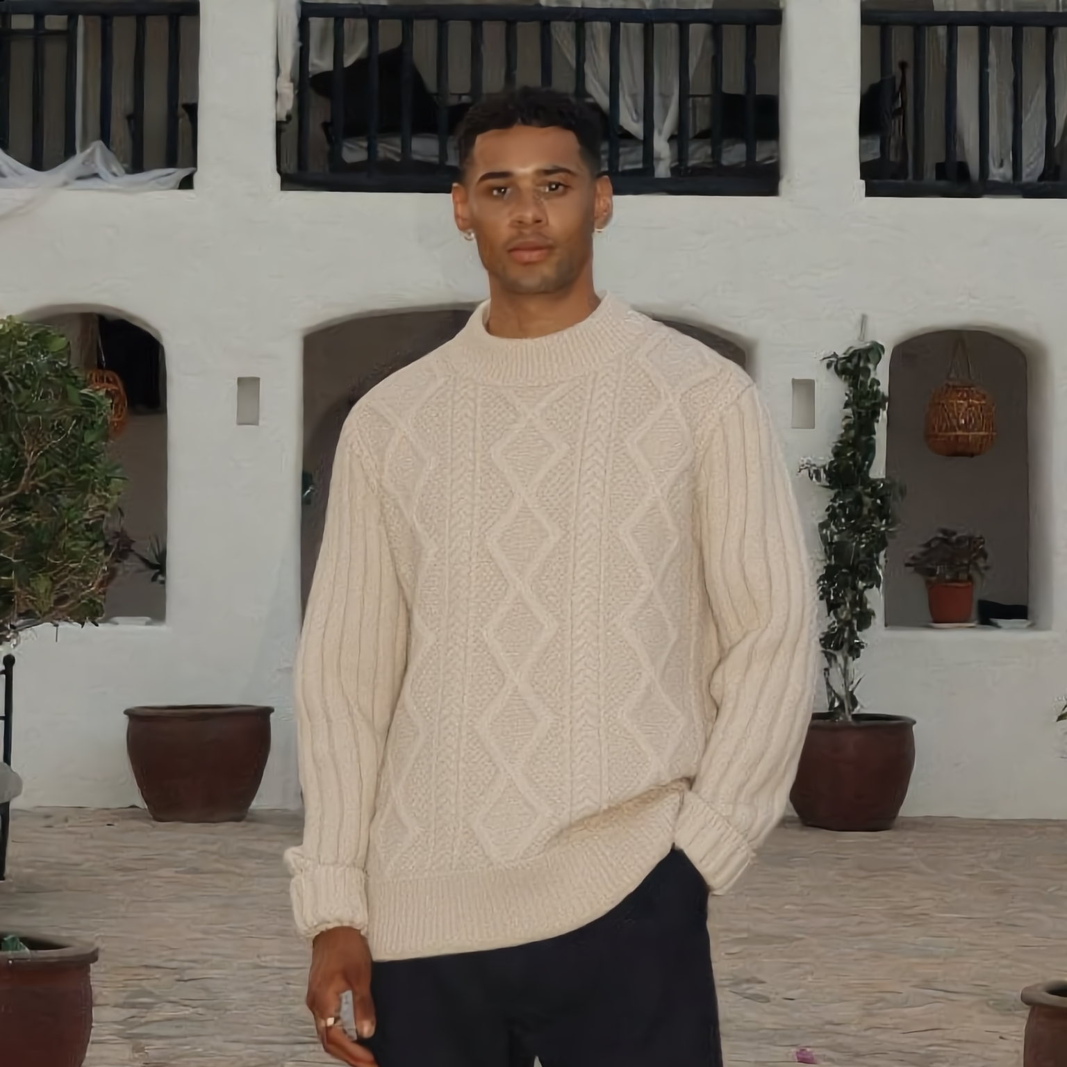 Man wearing a beige cable knit sweater and dark pants standing on a stone path.